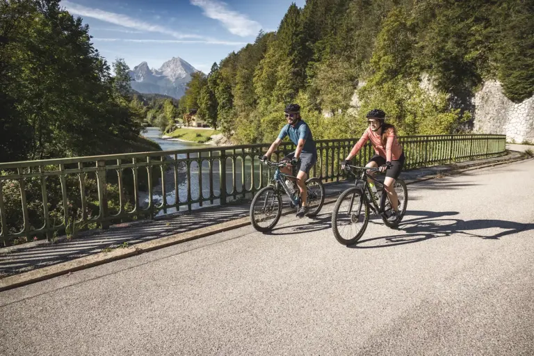 Zwei Radfahrer auf einer Brücke über die Ache vor Bergkulisse